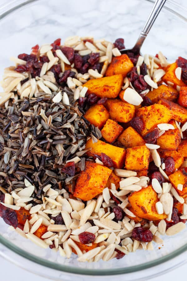 Roasted butternut squash, wild rice, cranberries, and slivered almonds in glass bowl with spoon.