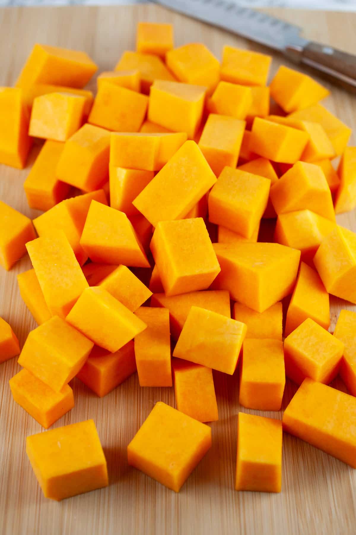 Diced butternut squash chunks on wooden cutting board with knife.