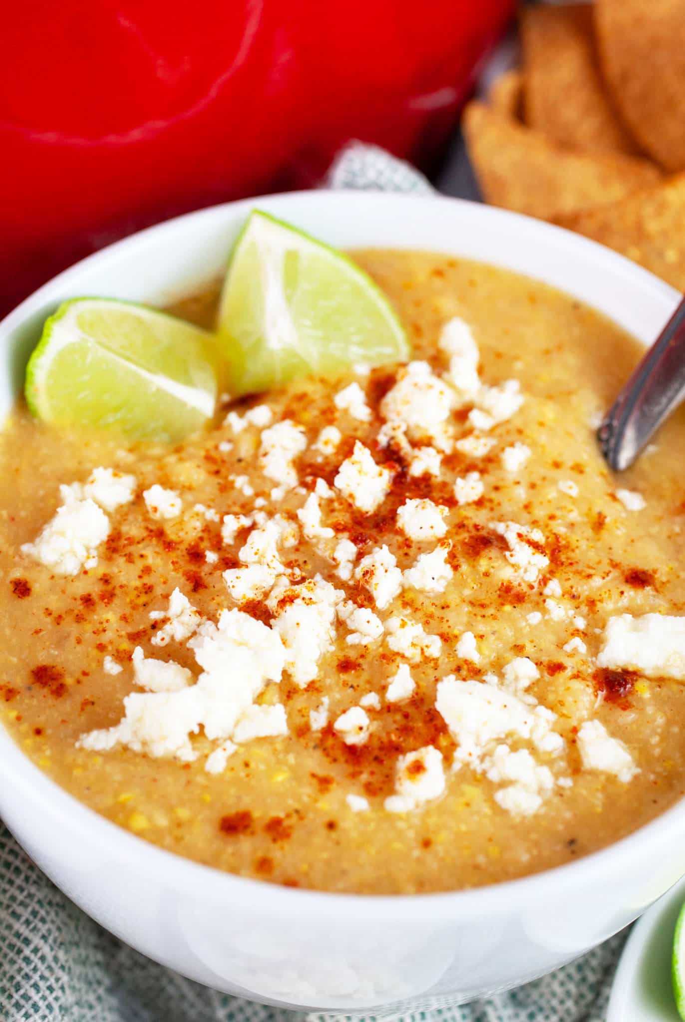 Southwest sweet corn cauliflower soup garnished with chili powder, queso fresco, and lime wedges in white bowl with spoon.