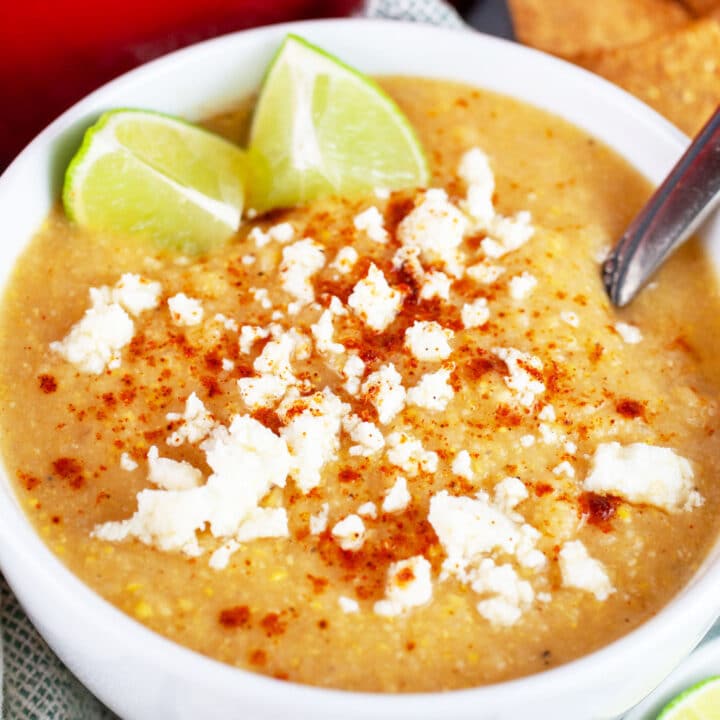 Southwest corn cauliflower soup garnished with queso fresco, chili powder, and lime wedges in white bowl with spoon.