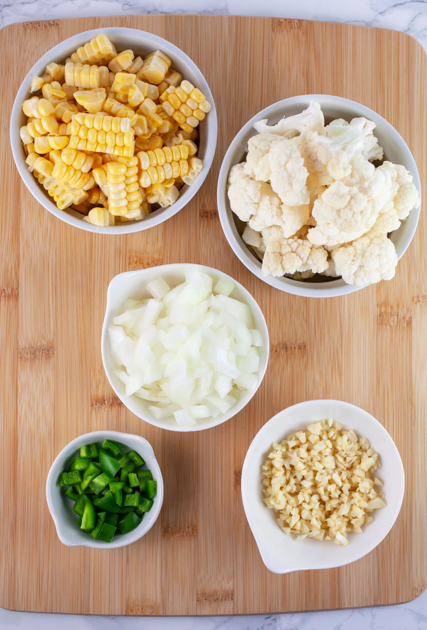 Minced garlic, onions, jalapeno pepper, corn kernels, and cauliflower florets in small white bowls on wooden cutting board.