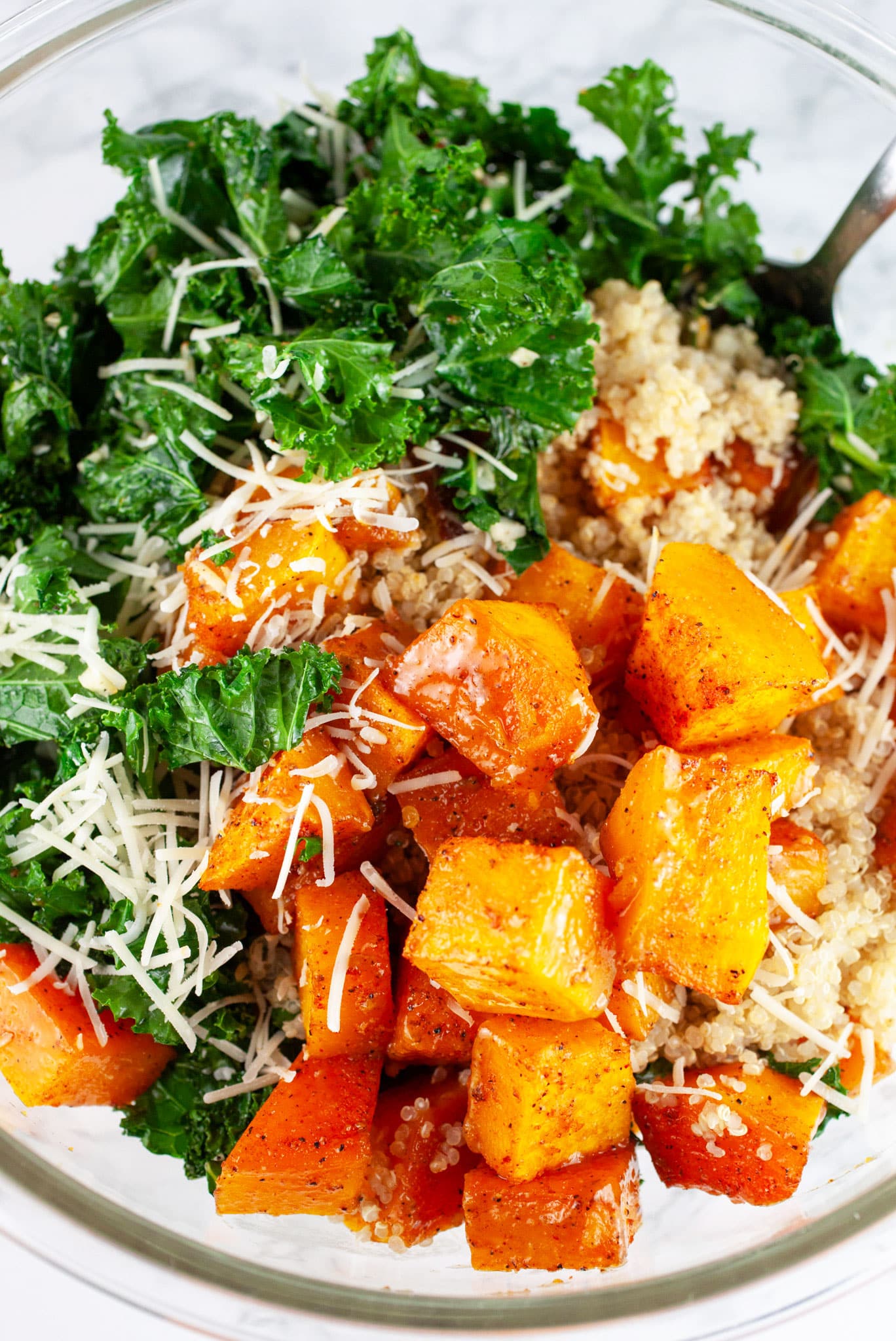 Roasted butternut squash, sauteed kale, quinoa, and Parmesan cheese in large glass bowl with spoon.