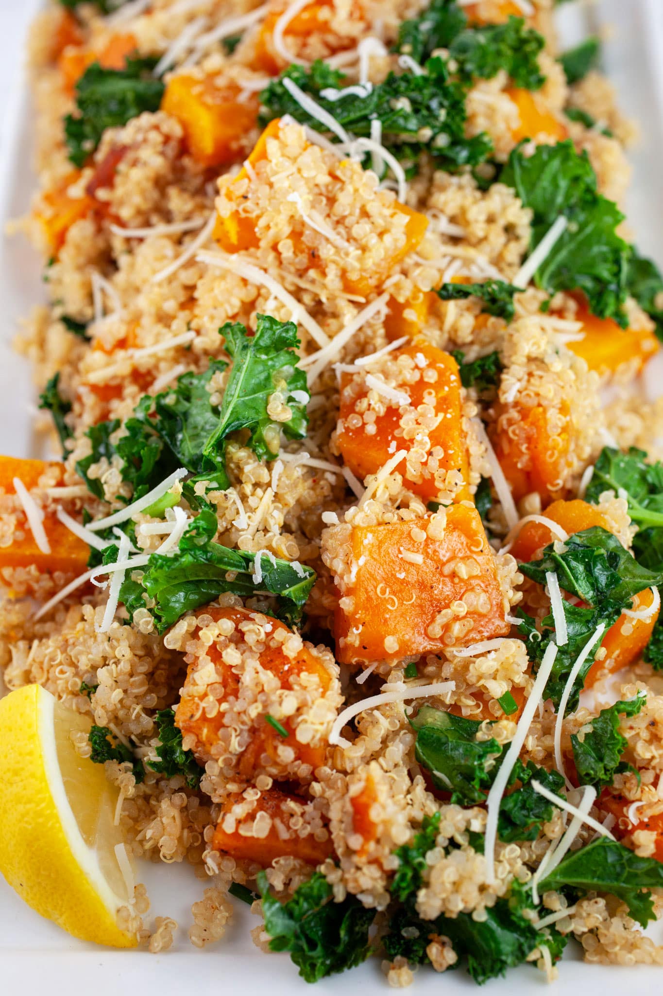Roasted butternut squash quinoa kale salad on white platter garnished with Parmesan cheese and lemon wedge.