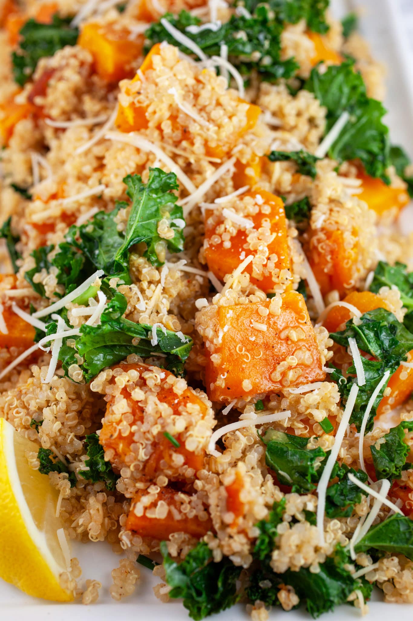 Butternut squash quinoa kale salad on white platter garnished with Parmesan cheese and lemon wedge.