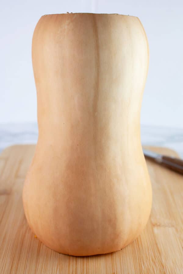 Butternut squash with top and bottom cut off on wooden cutting board with knife.