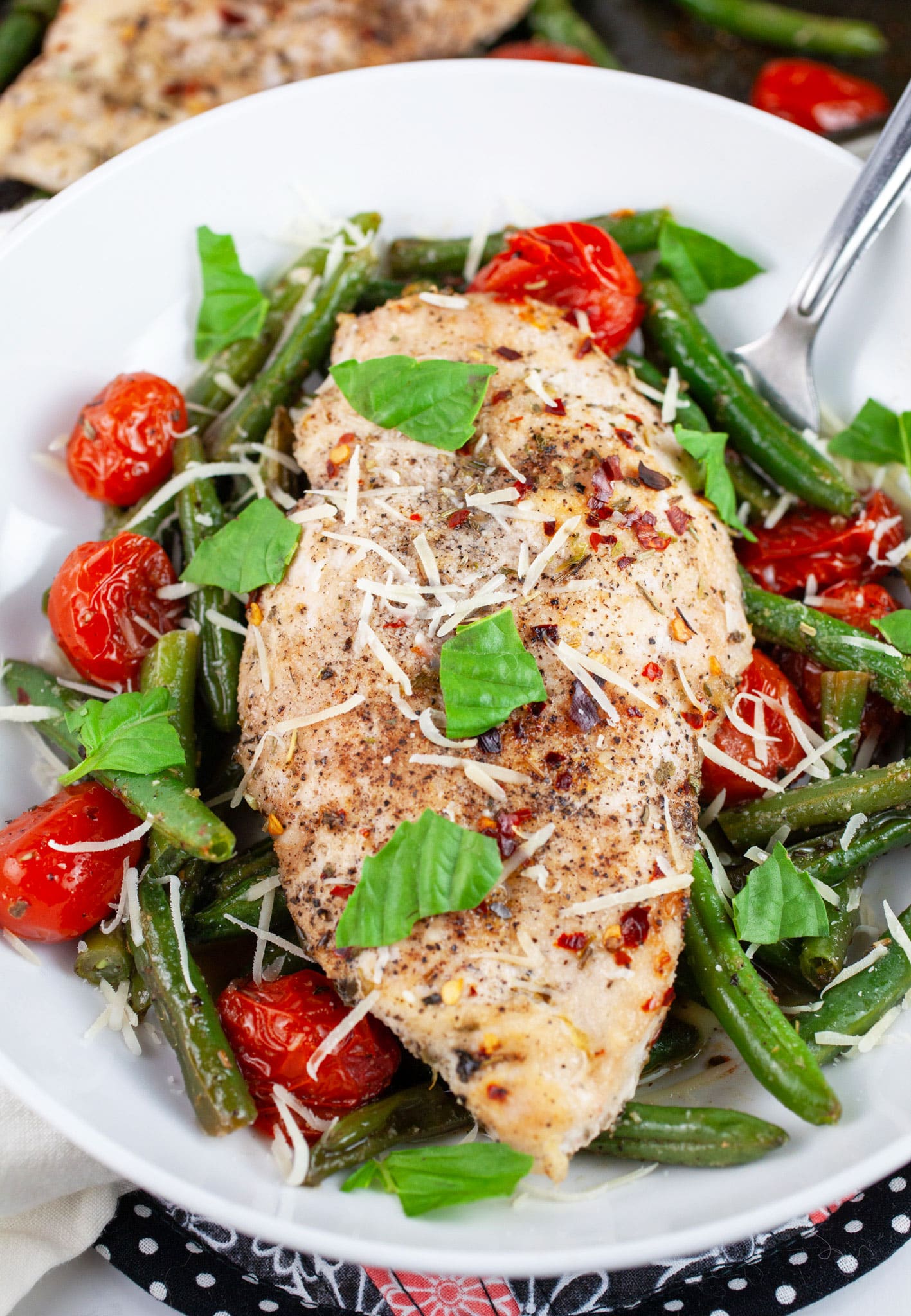 Italian chicken with roasted green beans and tomatoes garnished with Parmesan cheese and fresh basil in white bowl with fork.