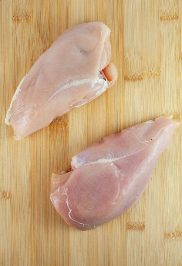 Whole raw boneless, skinless chicken breasts on wooden cutting board.
