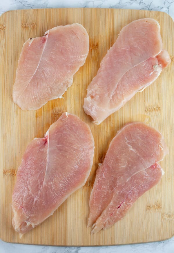 Raw chicken breasts cut in half lengthwise on wooden cutting board.