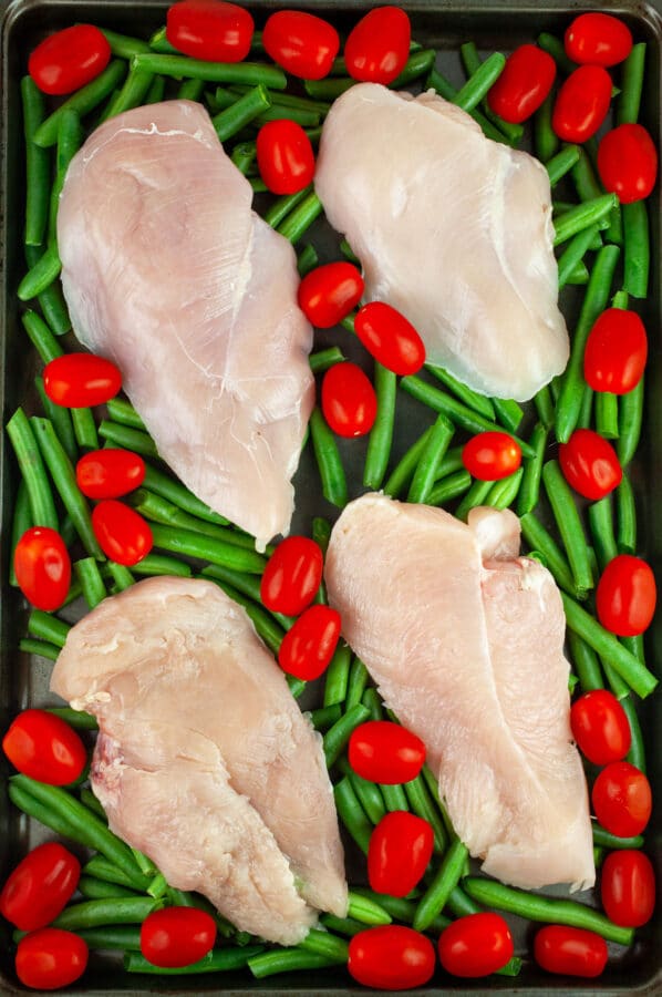 Raw chicken breasts, green beans, and grape tomatoes on baking sheet.