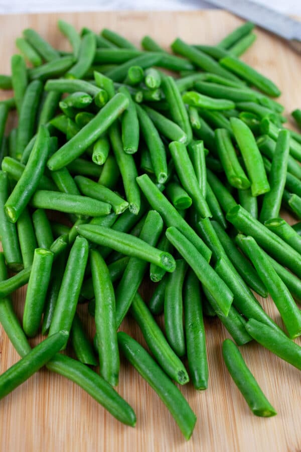 Fresh green beans cut into halves on wooden cutting board with knife.