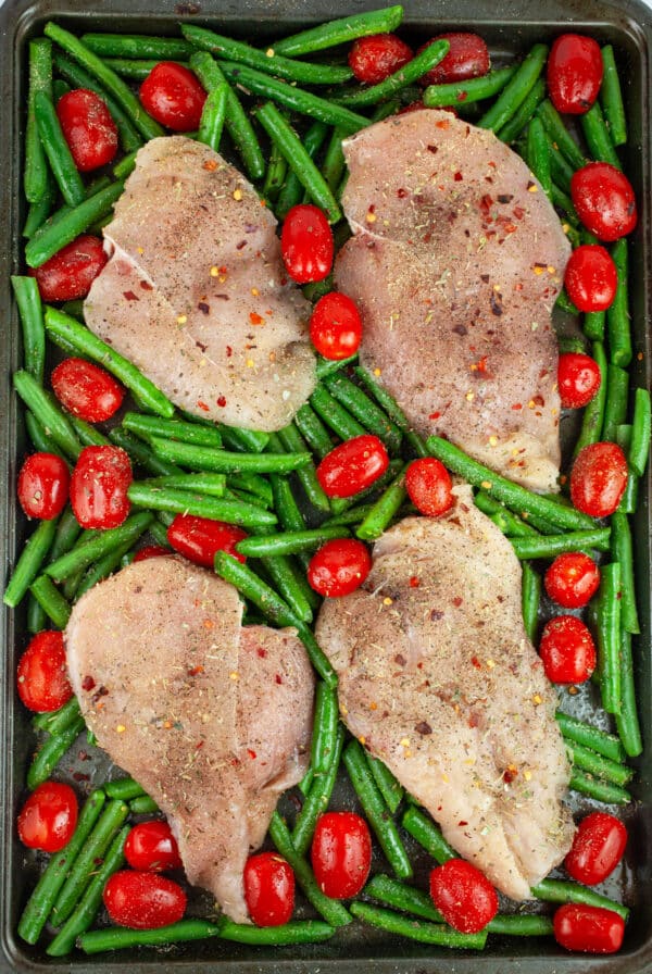 Uncooked, seasoned chicken breasts, green beans, and grape tomatoes on baking sheet.