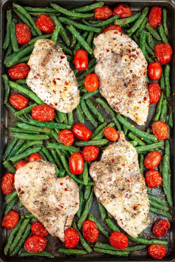 Cooked chicken breasts, green beans, and grape tomatoes on baking sheet.