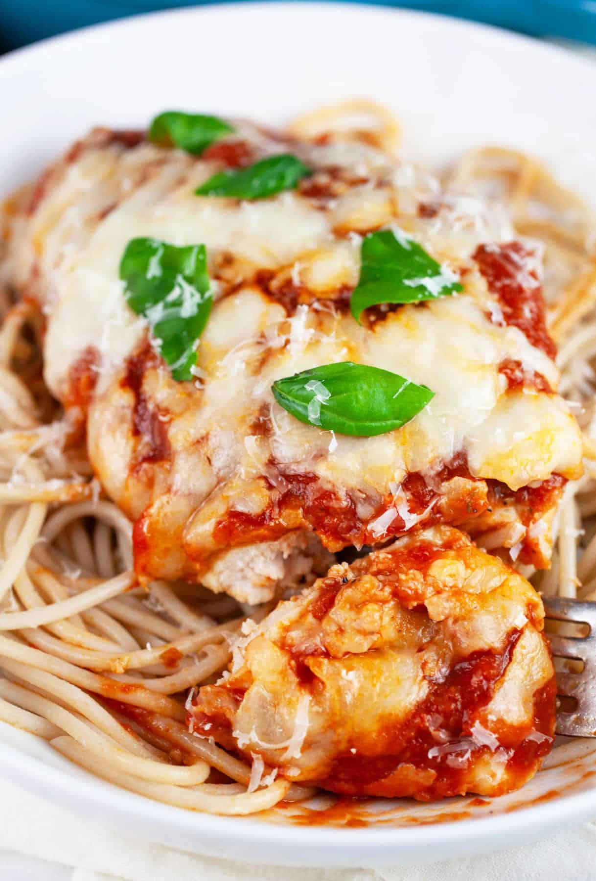 Chicken Parmesan with spaghetti garnished with fresh basil in white bowl with fork.