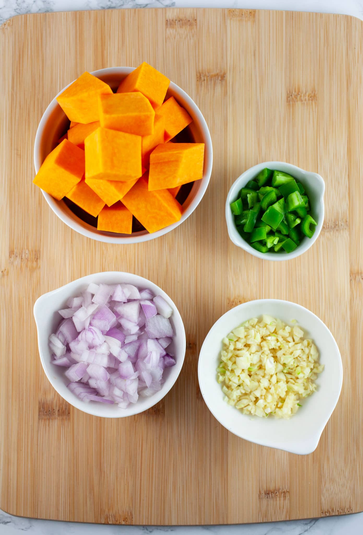 Minced garlic, onion, jalapeno pepper, and diced butternut squash in small white bowls on wooden cutting board.