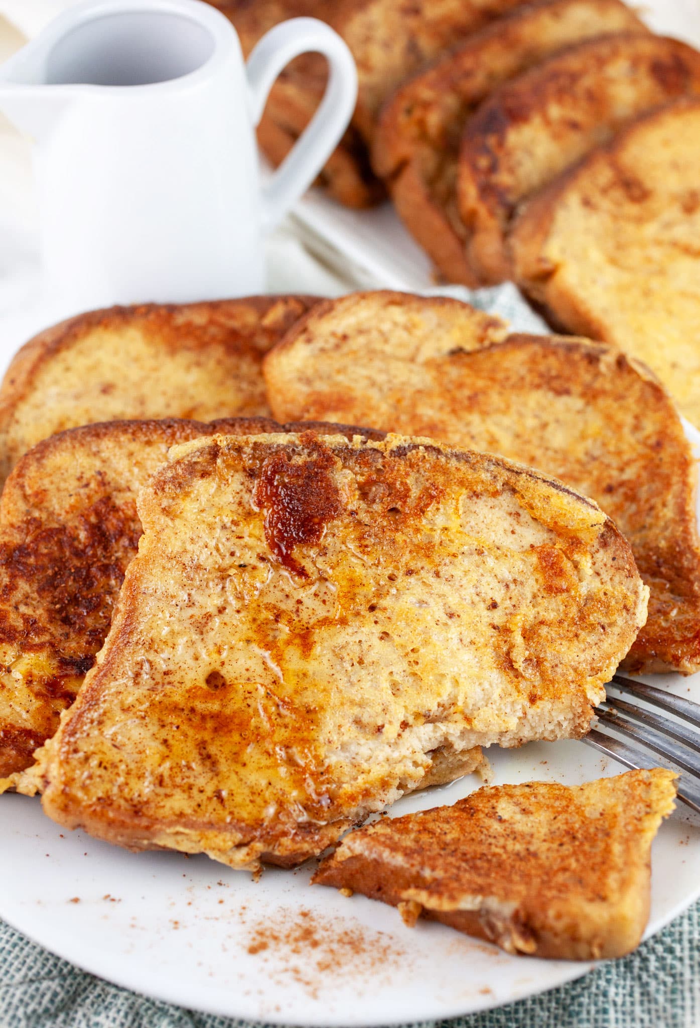 Eggnog French toast cut with fork on white plate next to container of maple syrup.