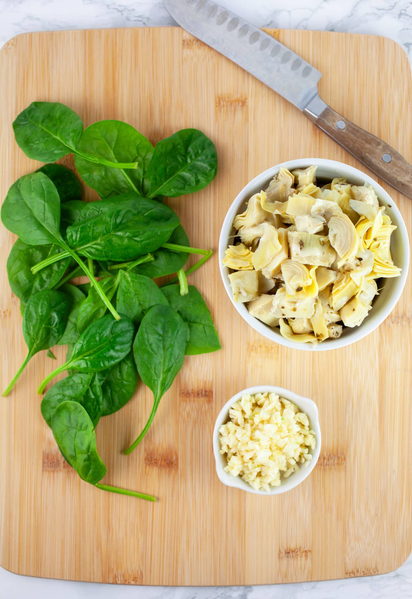 Minced garlic, chopped marinated artichoke hearts, and fresh spinach with knife on wooden cutting board.