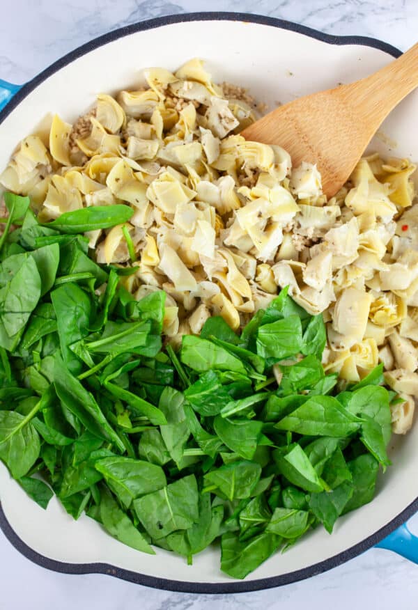 Chopped marinated artichoke hearts and fresh spinach in skillet with wooden spatula.