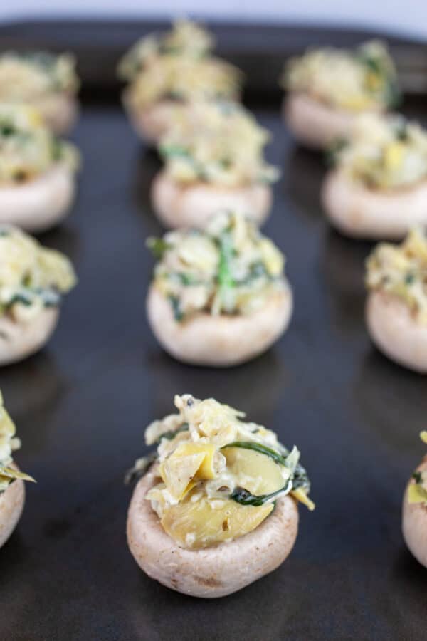 Uncooked spinach artichoke stuffed mushrooms on baking sheet.
