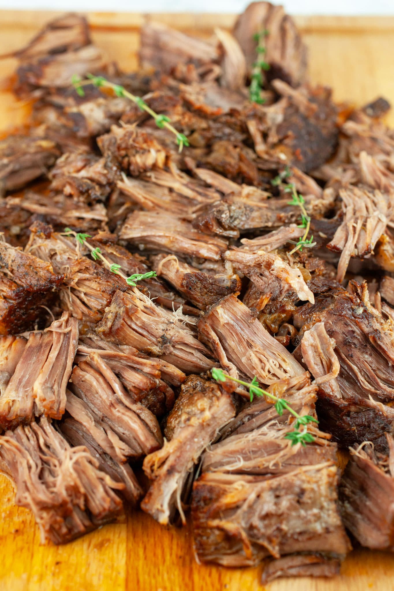 Shredded, cooked beef pot roast on wooden cutting board garnished with fresh thyme.