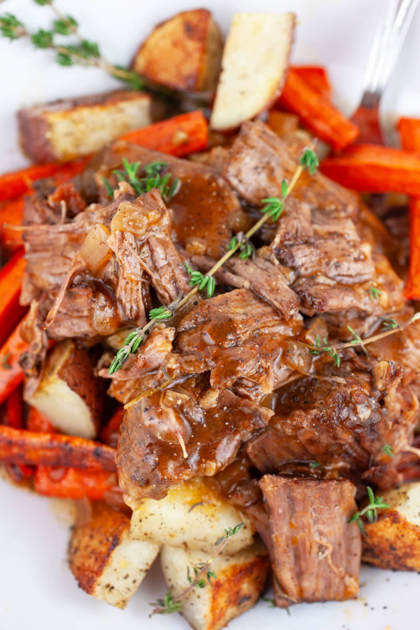 Pot roast with roasted potatoes and carrots garnished with fresh thyme in white bowl with fork.