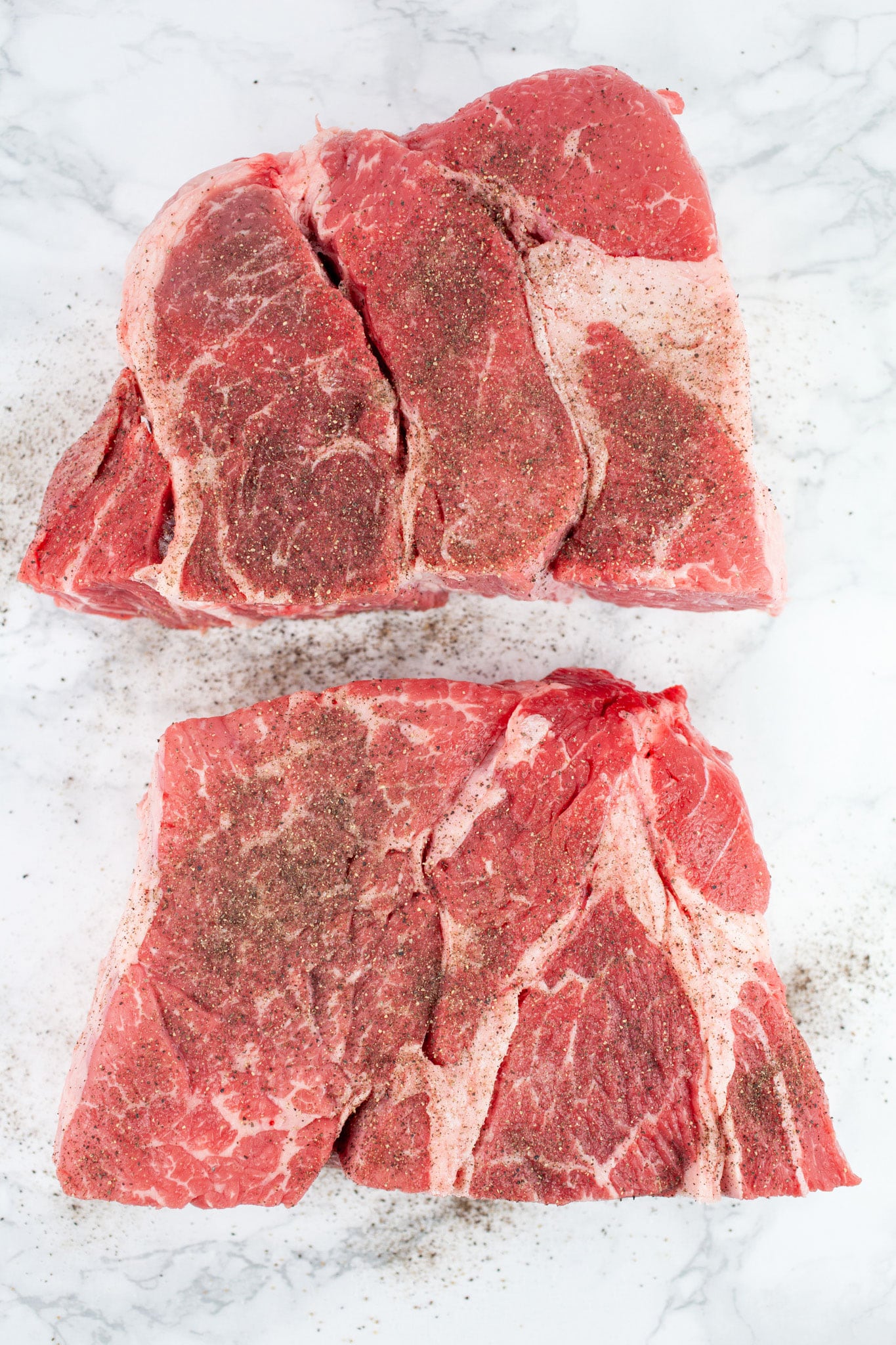 Raw boneless beef chuck roast cut in half and seasoned with kosher salt and pepper on white surface.