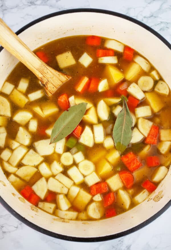 Diced vegetables with broth and bay leaves in Dutch oven with wooden spoon.