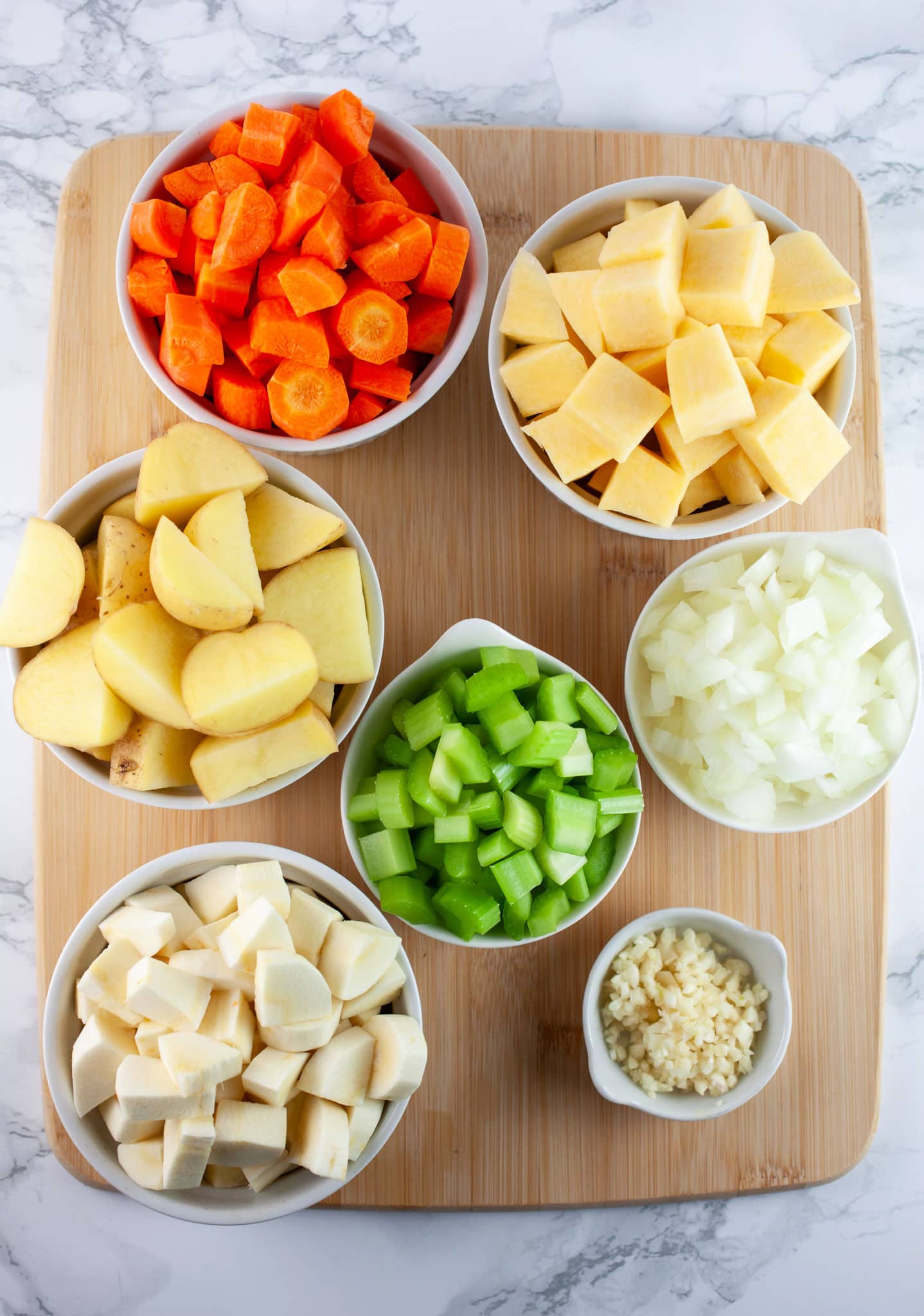 Minced garlic, onions, and celery and chopped parsnips, rutabaga, carrots, and potatoes in white bowls on wooden cutting board.