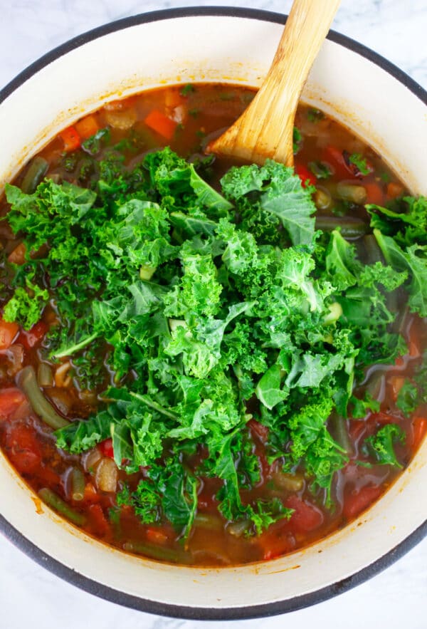 Chopped kale added to vegetable soup in Dutch oven with wooden spoon.