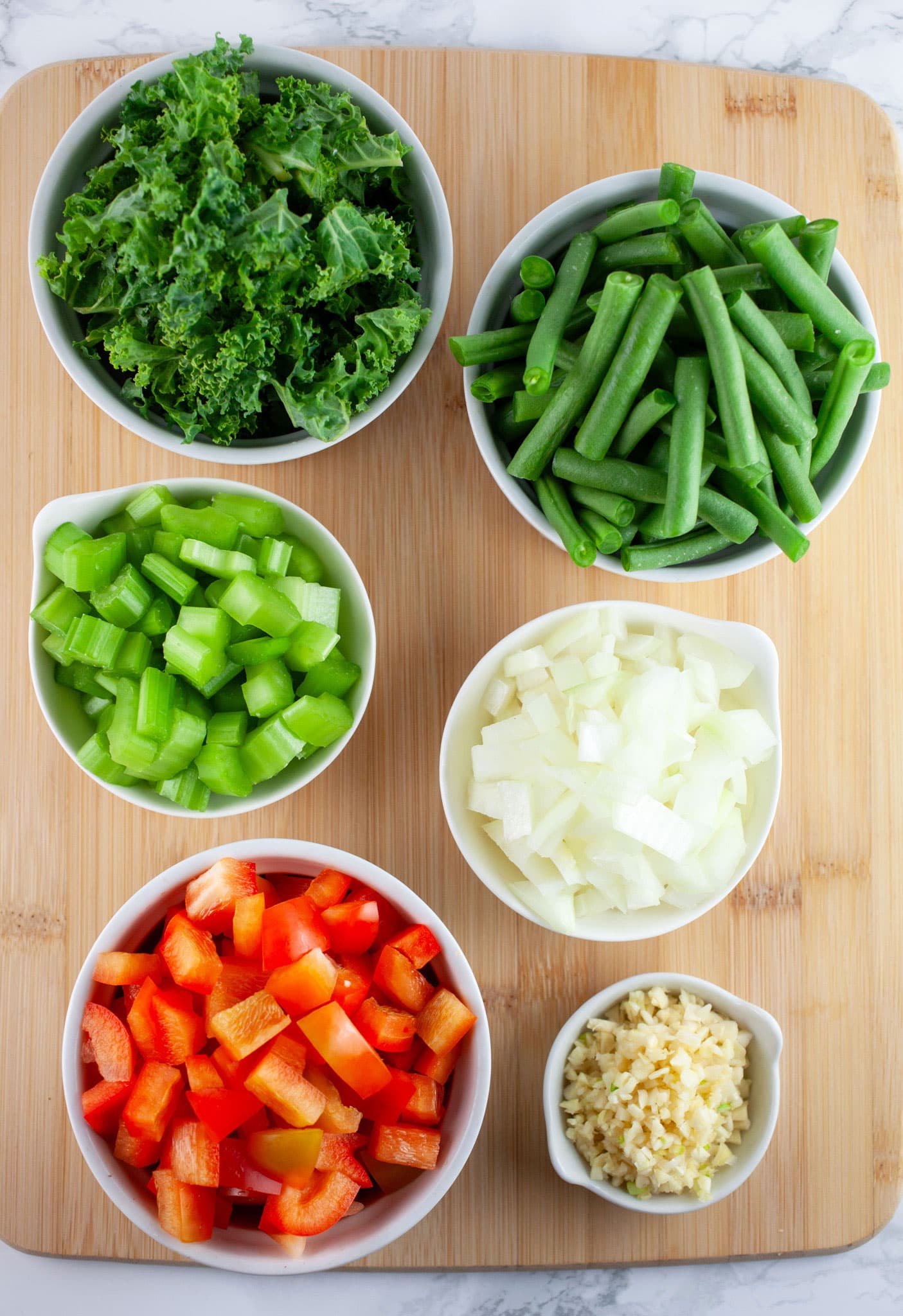 Minced garlic, onions, celery, red bell peppers, green beans, and kale in small white bowls on wooden cutting board.