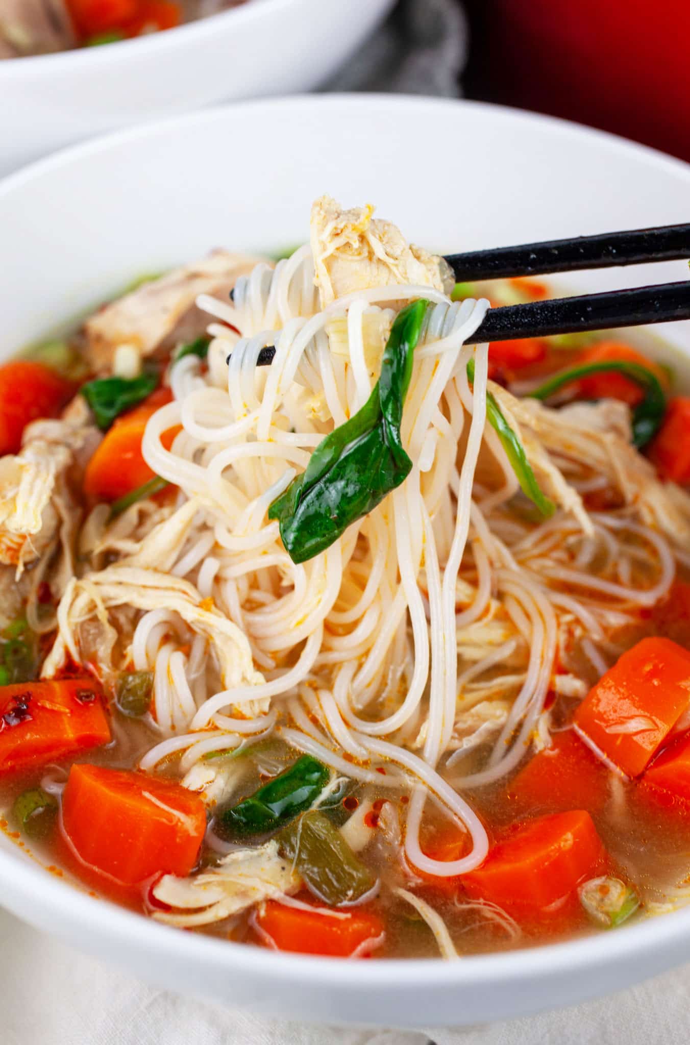 Chopsticks lifting rice noodles from bowl of Asian chicken soup.