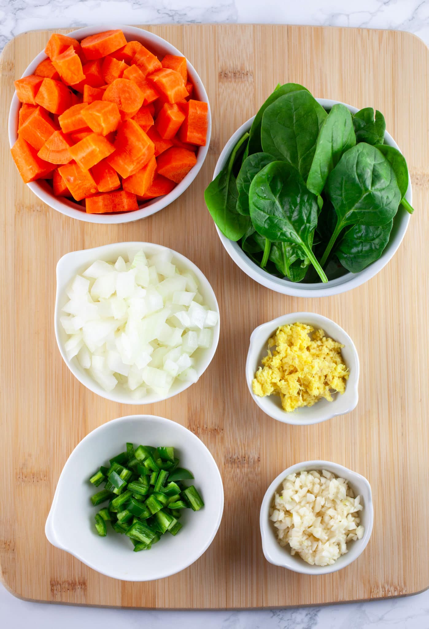 Minced garlic, onions, ginger, and jalapeno, chopped carrots, and spinach in small white bowls on wooden cutting board.