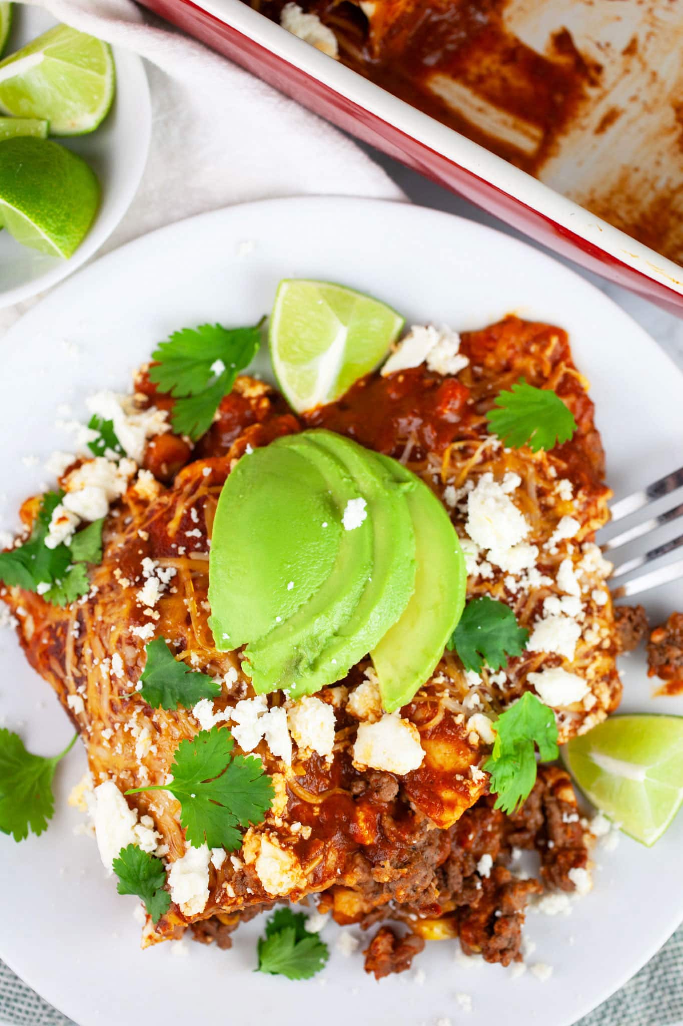Ground beef enchiladas with Queso fresco, sliced avocadoes, cilantro, and lime wedges on white plate with fork.