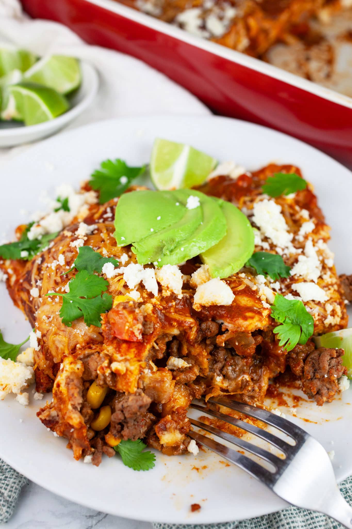 Ground beef enchiladas on white plate with fork garnished with Queso fresco, cilantro, sliced avocadoes, and limes.