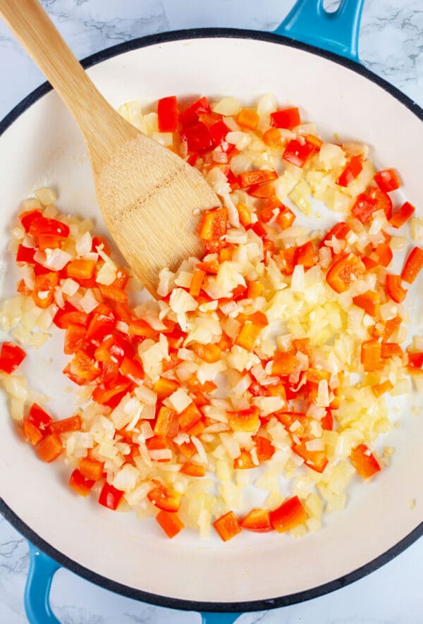 Minced garlic, onions, and red bell peppers sauteed in skillet with wooden spoon.