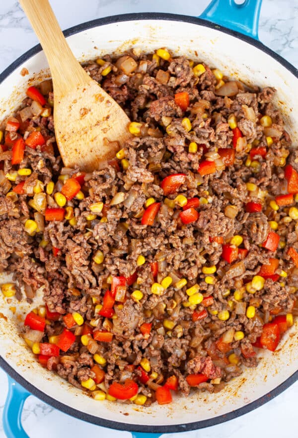 Ground beef, red bell peppers, and corn sauteed in skillet with wooden spoon.