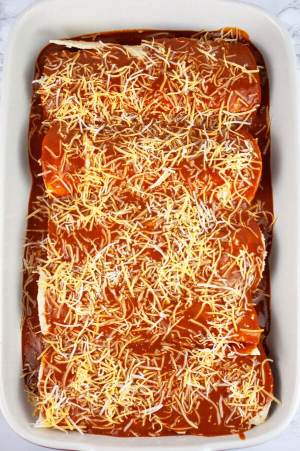 Filled tortillas covered with red enchilada sauce and shredded cheese in casserole dish.