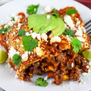 Ground beef enchiladas garnished with cilantro, avocado, and Queso fresco on white plate with fork.