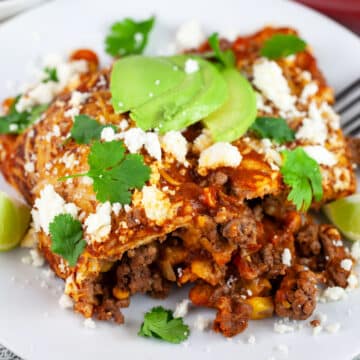 Ground beef enchiladas garnished with cilantro, avocado, and Queso fresco on white plate with fork.