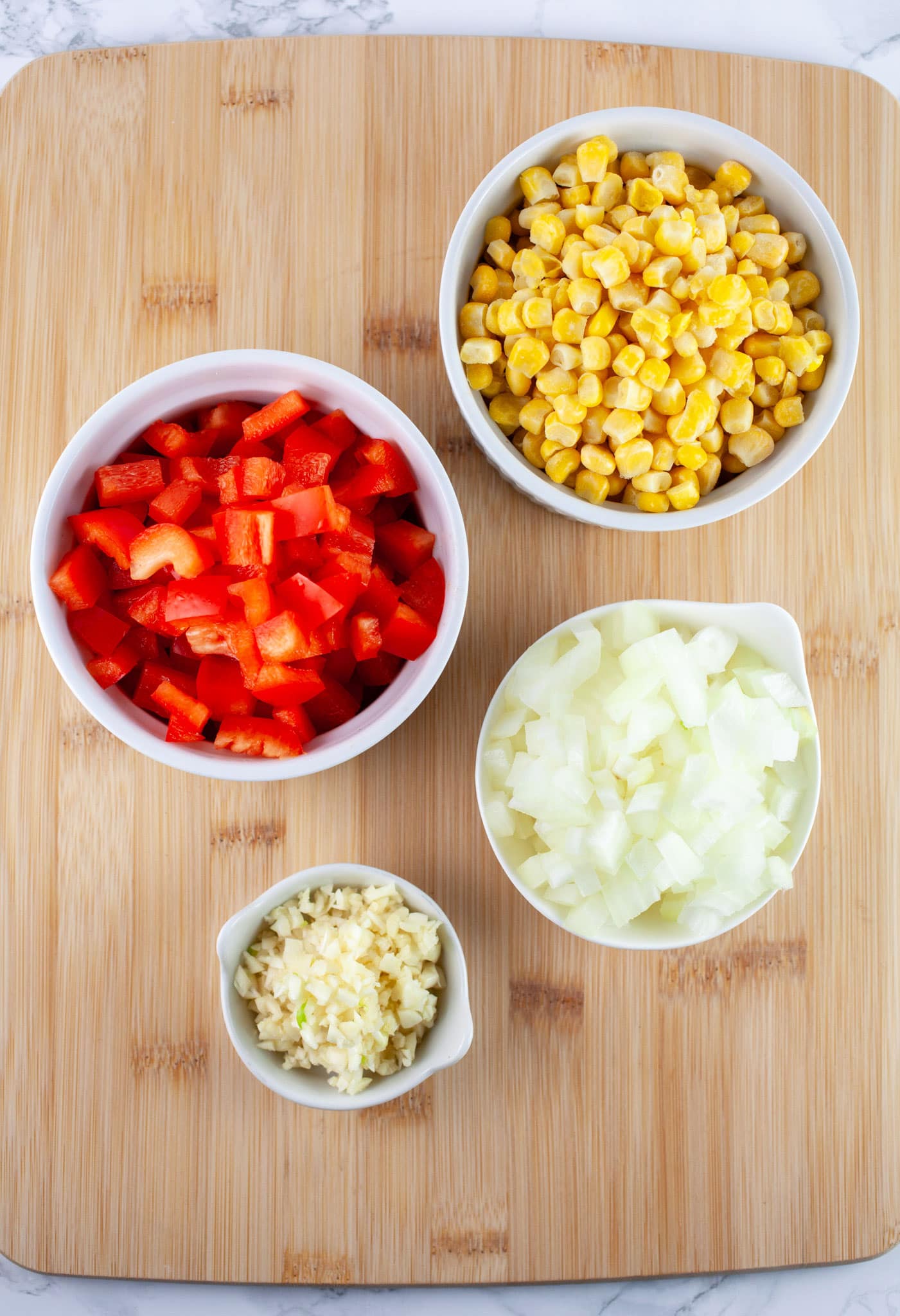 Minced garlic and onions, diced red bell peppers, and frozen corn in small white bowls on wooden cutting board.