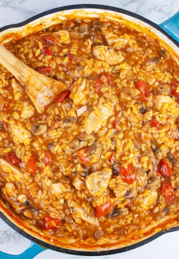 Cooked chicken, mushroom, and tomato pasta in skillet with wooden spoon.