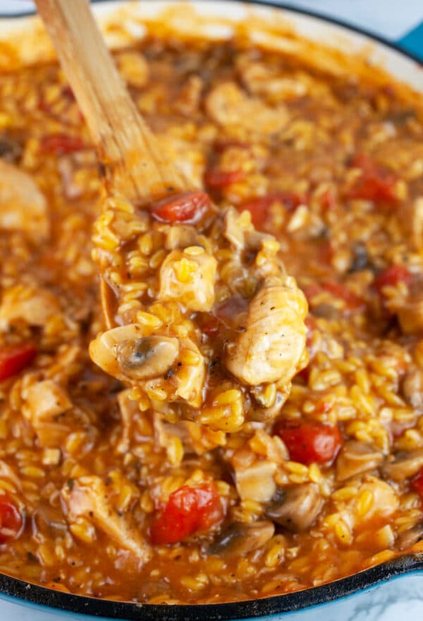 Scoop of chicken mushroom tomato pasta lifted from skillet on wooden spoon.