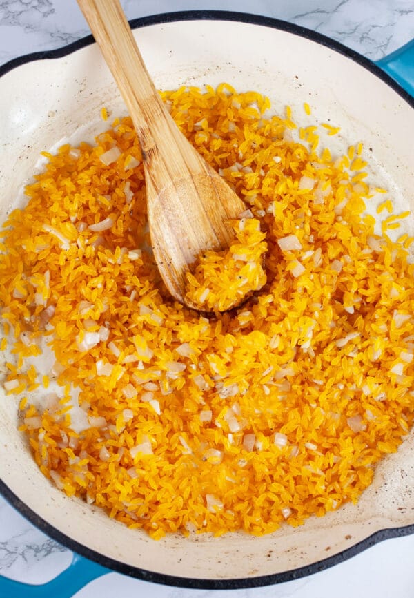 Minced garlic, shallots, and orzo pasta sauteed in skillet with wooden spoon.
