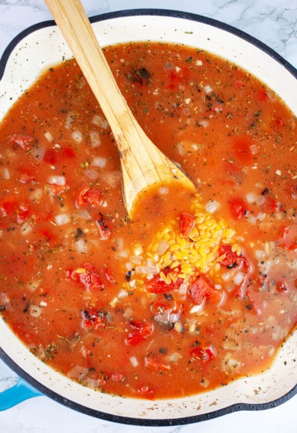 Uncooked orzo pasta with fire roasted tomatoes in skillet with wooden spoon.