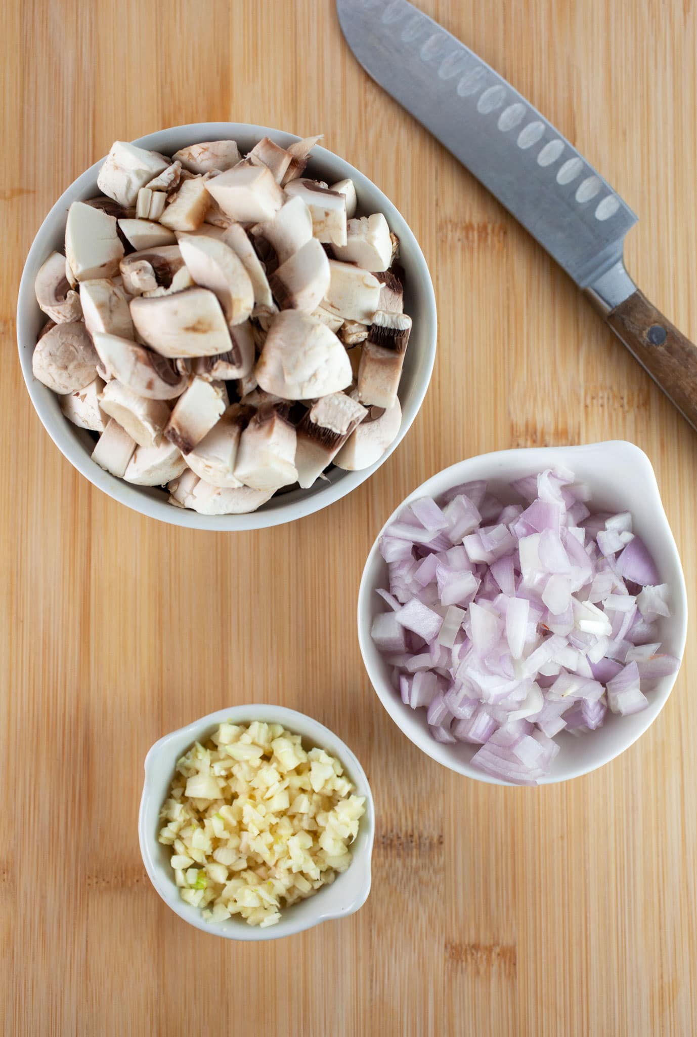 Minced garlic, shallots, and chopped mushrooms in small white bowls on wooden cutting board with knife.