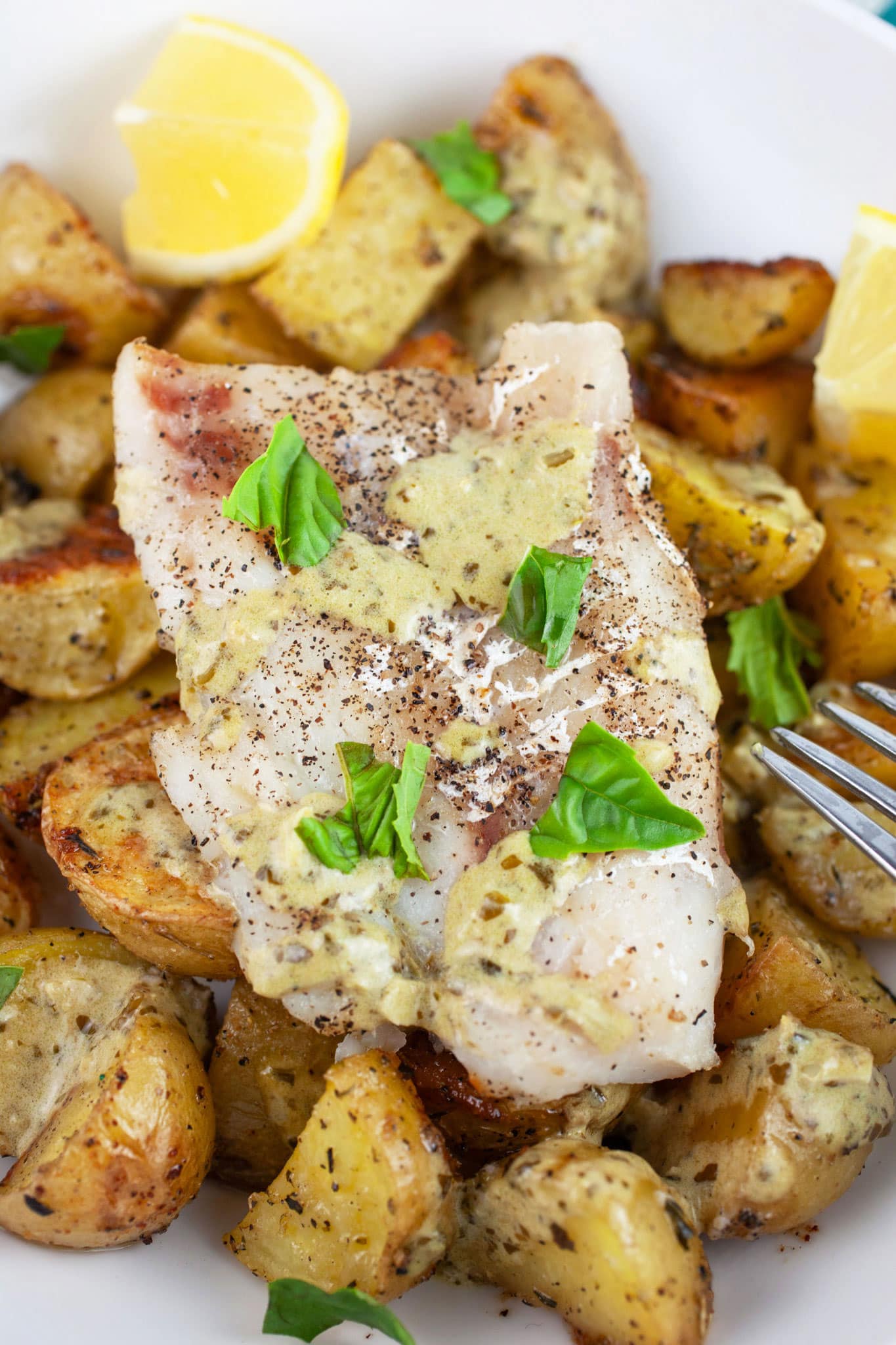 Baked cod and potatoes in white bowl with fork garnished with pesto sauce, fresh basil, and lemon wedges.