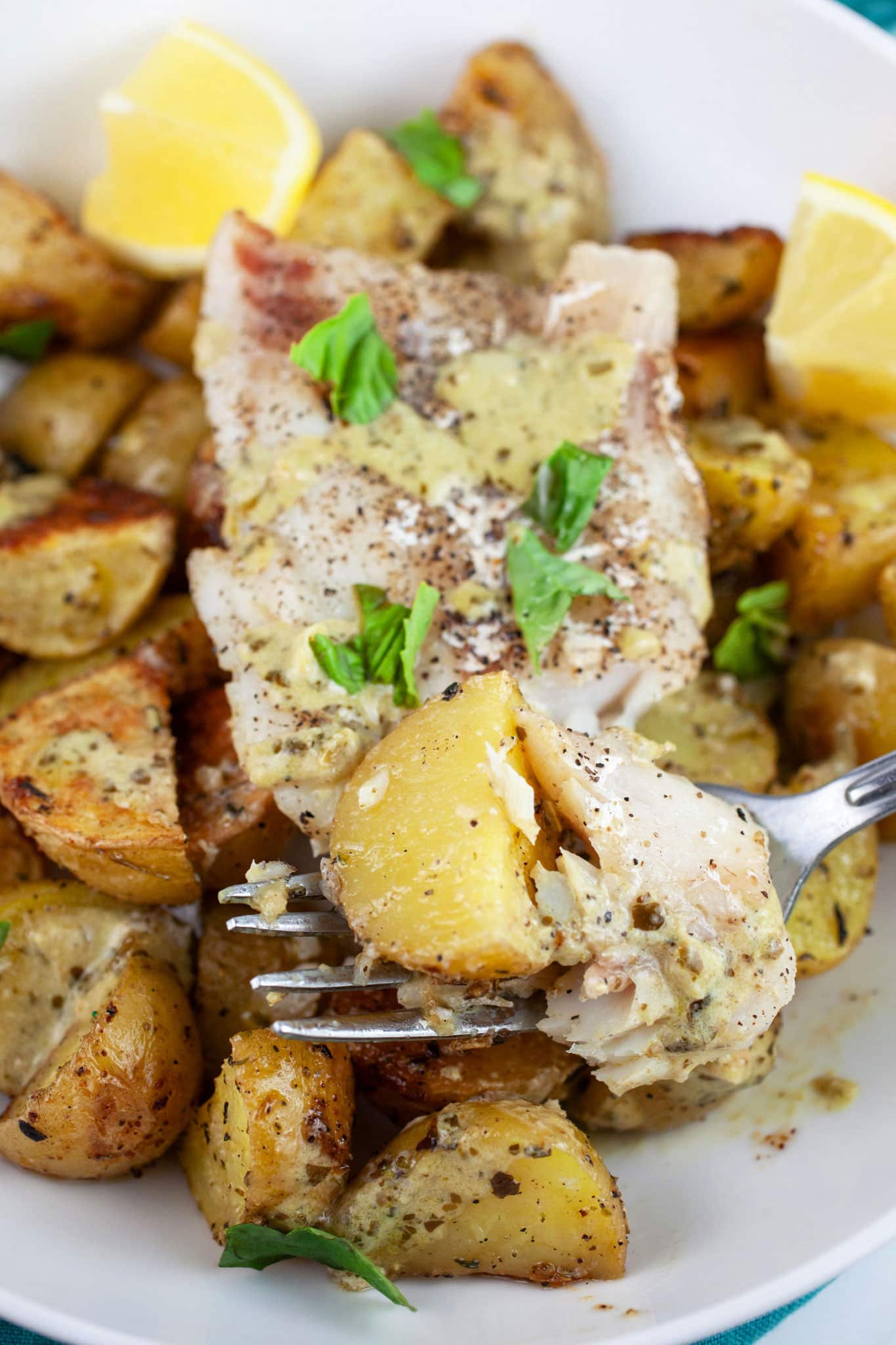 Pesto baked cod and potatoes in white bowl with fork garnished with fresh basil and lemon wedges.