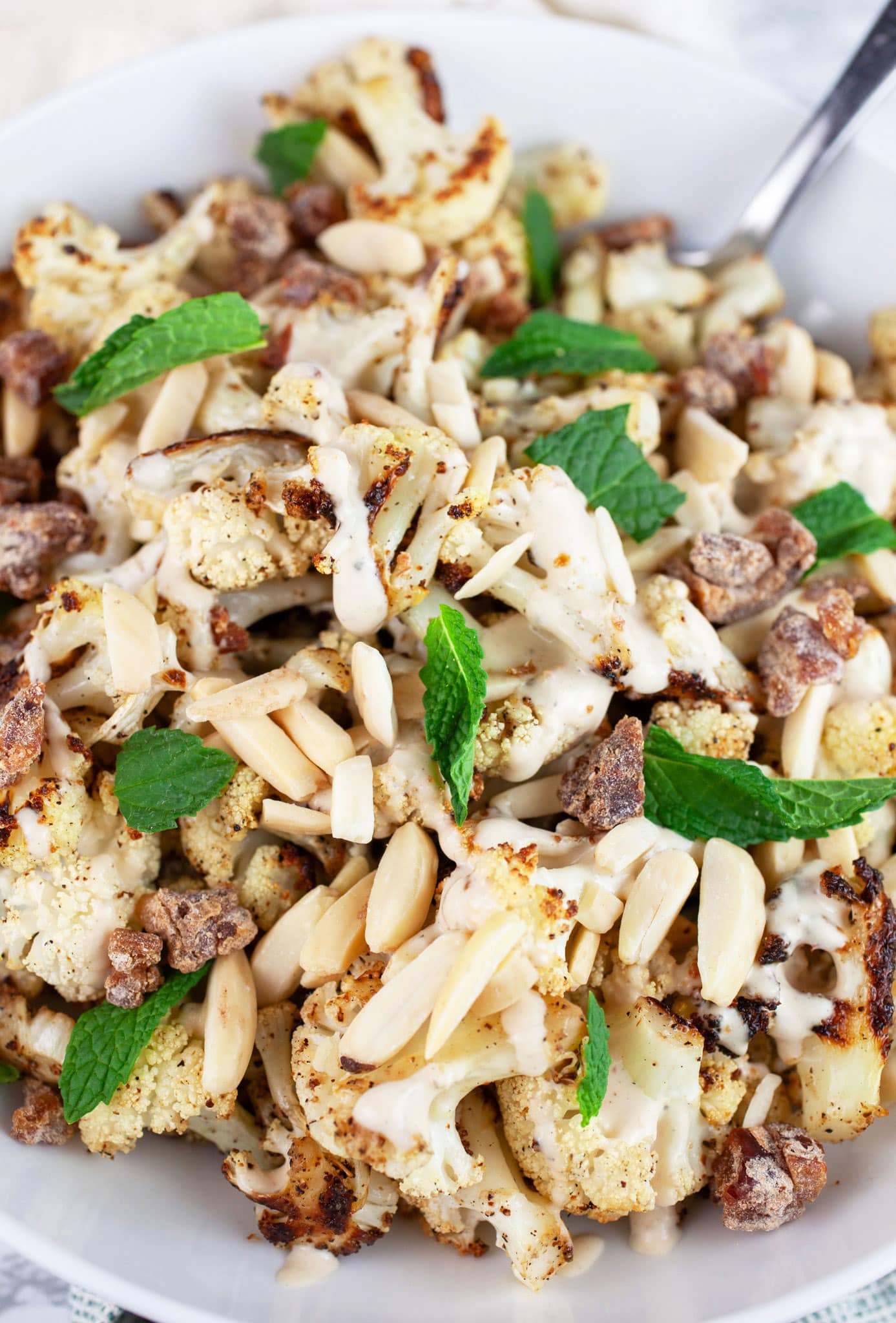Roasted Mediterranean cauliflower in white bowl with tahini sauce, slivered almonds, dates, and fresh mint.