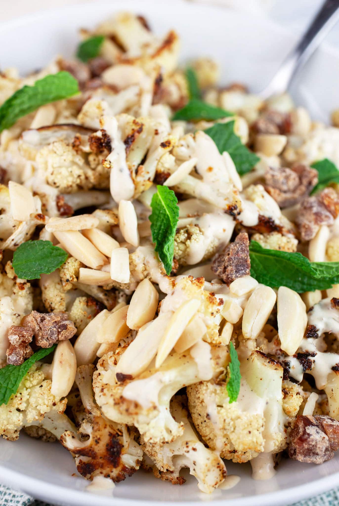 Mediterranean roasted cauliflower with lemon tahini sauce, slivered almonds, chopped dates, and fresh mint in white bowl with fork.