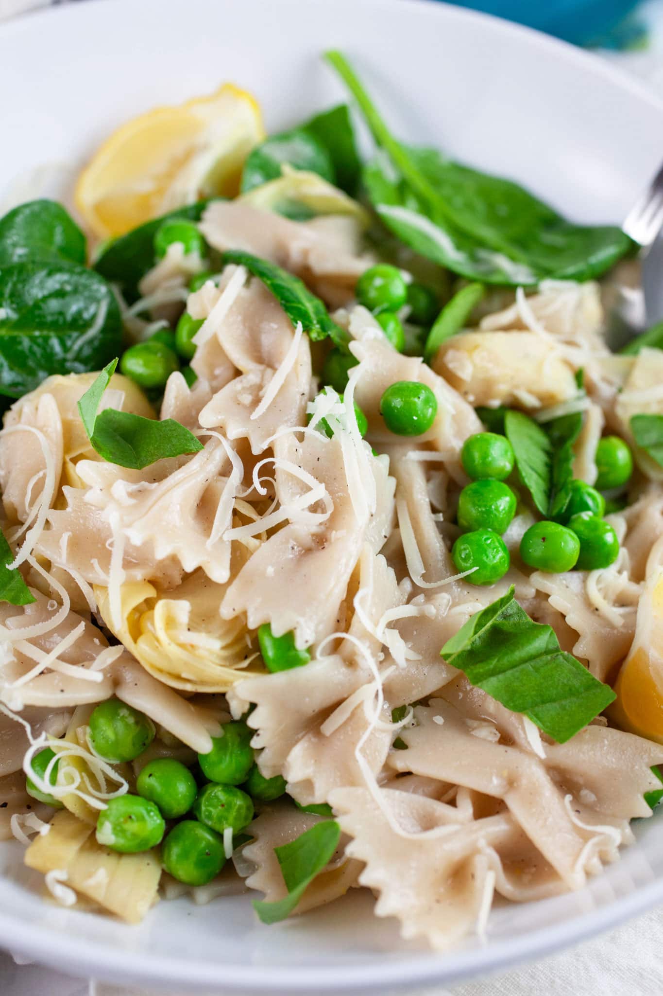 Spring vegetable pasta in white bowl with fork garnished with Parmesan cheese, lemon wedges and basil.
