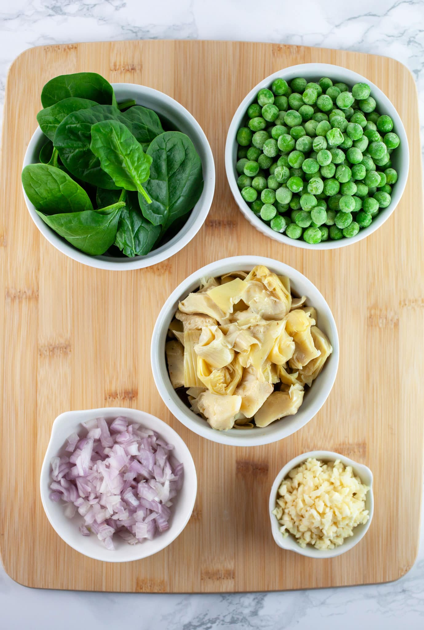 Minced garlic and shallots, marinated artichoke hearts, frozen peas, and fresh spinach in small white bowls on wooden cutting board.